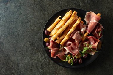 Siyah tabakta ekmek çubuğu, zeytin ve biberiyeli jambon. Taş bir masada geleneksel İtalyan atıştırmalıkları. Üst görünüm.