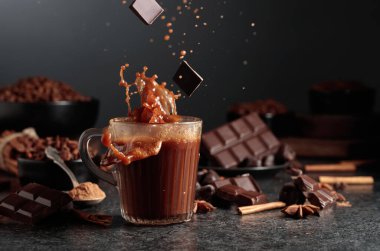 Siyah mutfak masasının üzerinde malzemeler olan bir bardak sıcak çikolata. Kakao parçacıkları bir bardak kakao içeceğinin içine düşer ve güzel bir su sıçratır..