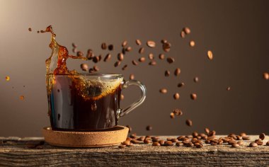 Eski bir tahtanın üzerinde cam bardakta sade kahve. Kavrulmuş kahve çekirdekleri hareket halinde. Boşluğu kopyala.