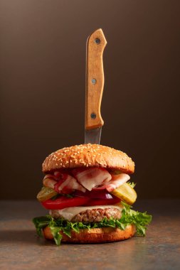 Kahverengi zemin üzerinde bıçak olan taze bir burger. Domatesli hamburger, soğan, korunmuş salatalık, salata, peynir, sığır eti ve pastırma..