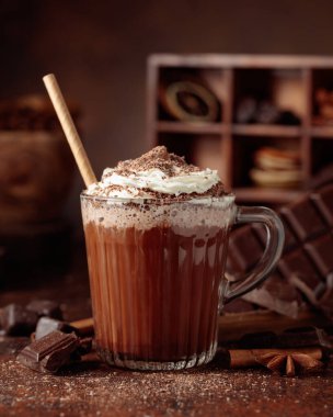 Üzerine krem şanti ve çikolata parçacıkları serpiştirilmiş sıcak çikolata. Eskimiş kahverengi arka planda yer alan malzemeler içeren bir çikolata içeceği..