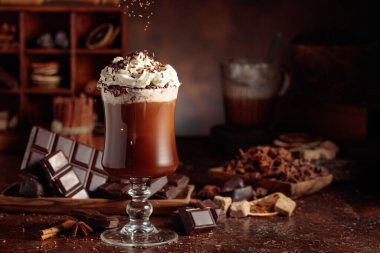 Üzerine krem şanti ve çikolata parçacıkları serpiştirilmiş sıcak çikolata. Eskimiş kahverengi arka planda yer alan malzemeler içeren bir çikolata içeceği..