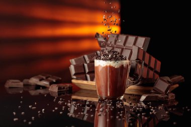 Çikolata kırıntıları serpiştirilmiş kremalı sıcak çikolata. Arka planda kırılmış acı çikolatalar. Siyah yansıtıcı arkaplan.