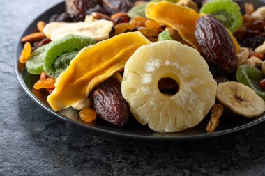 Kurumuş tropikal meyveler, fındıklar ve kuru üzümler siyah bir taş masada..