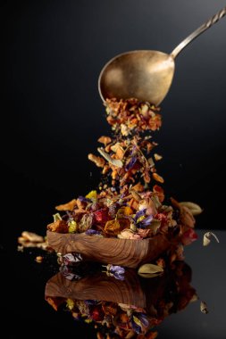 Çeşitli kurutulmuş otlar ve baharatlar ahşap bir kaba dökülür. Siyah yansıtıcı arkaplan.