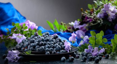 Siyah tabakta taze yaban mersini. Böğürtlenlerle, renkli bezelyelerle ve koyu mavi arka planda çayır otlarıyla dolu bir yaz..