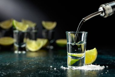 Tekila bardağa dökülür. Koyu mavi bir masada tekila, limon dilimleri ve deniz tuzu..