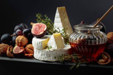 Taze incir, üzüm, gevrek ceviz, serpiştirilmiş bal ve mis kokulu kekik eşliğinde nefis bir kremalı Camembert peyniri muhteşem bir görsel cazibe için siyah bir arkaplanda zarif bir şekilde sunuluyor..