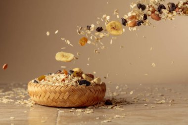 Müsli bej bir arka planda hareket halinde. Yulaf gevreği, fındık, kuru üzüm ve kuru muz dilimleri bir kaseye düşer. Boşluğu kopyala.