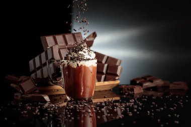 Çikolata kırıntıları serpiştirilmiş kremalı sıcak çikolata. Arka planda kırılmış acı çikolatalar. Siyah yansıtıcı arkaplan.