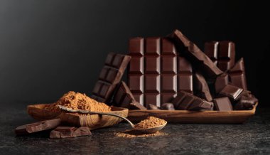 Tahta bir kase kakao tozunun yanında koyu çikolata kırıntıları. Siyah arka plan. Kakao tozlu bir kaşığa odaklan. Boşluğu kopyala.