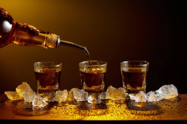 Viski şişeden cama doluyor. Viski ve buz siyah bir masada. Sarı ışık güzel gölgeler yaratır. Boşluğu kopyala.