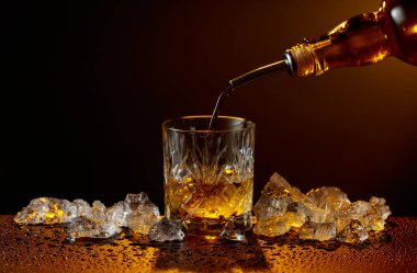 Viski şişeden cama doluyor. Viski ve buz, siyah ıslak bir masada. Sarı ışık, boşluğu kopyala.