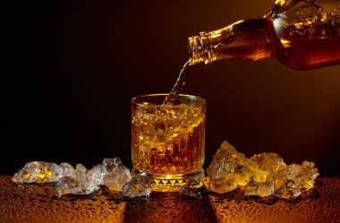 Viski şişeden cama doluyor. Viski ve buz, siyah ıslak bir masada. Sarı ışık, boşluğu kopyala.