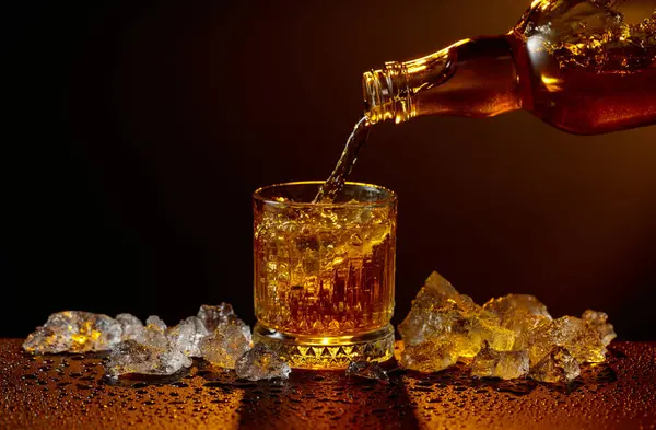Viski şişeden cama doluyor. Viski ve buz, siyah ıslak bir masada. Sarı ışık, boşluğu kopyala.