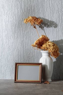 Boş resim çerçevesi ve gri duvarda kuru çiçekli beyaz vazo. Altın süslemeli ahşap çerçeve. Görüntüyü çerçeveye yerleştir. Boşluğu kopyala.