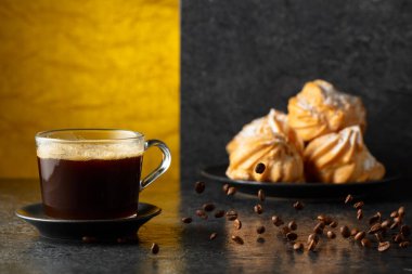 Cam bardakta sade kahve ve siyah masada hareketli kahve çekirdekleri. Arka plandaki kremalı keklerin içinde. Boşluğu kopyala.