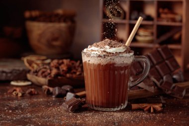 Üzerine krem şanti ve çikolata parçacıkları serpiştirilmiş sıcak çikolata. Eskimiş kahverengi arka planda yer alan malzemeler içeren bir çikolata içeceği..