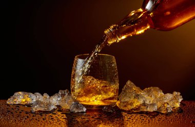 Viski şişeden cama doluyor. Viski ve buz, siyah ıslak bir masada. Sarı ışık, boşluğu kopyala.