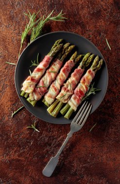 Domuz pastırmasına sarılı ızgara yeşil kuşkonmaz. Kahverengi arka planda siyah tabakta ev yapımı aperatif. Üst görünüm.