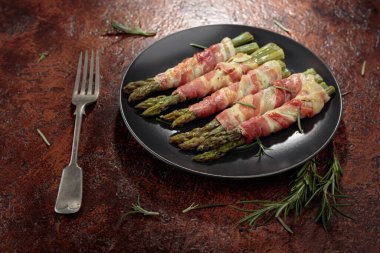 Izgara yeşil kuşkonmaz. Siyah tabakta domuz pastırması. Kahverengi bir arka planda..