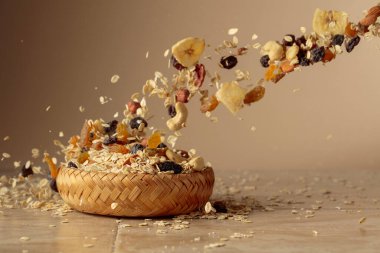 Müsli bej bir arka planda hareket halinde. Yulaf gevreği, fındık, kuru üzüm ve kuru muz dilimleri bir kaseye düşer. Boşluğu kopyala.