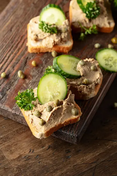 Eski bir kesme tahtasında ezmeli tost. Pate, taze salatalık, kapari ve maydanozlu sandviç..