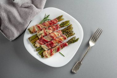 Domuz pastırmasına sarılı ızgara yeşil kuşkonmaz. Gri arka planda beyaz tabakta fotokopi alanı olan ev yapımı atıştırmalıklar. Üst görünüm.