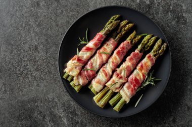 Domuz pastırmasına sarılı ızgara yeşil kuşkonmaz. Siyah tabakta ev yapımı atıştırmalık. Arka planda fotokopi alanı var. Üst görünüm.