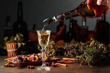 Sarı bitkisel likör ya da karışım eski bir şişeden bardağa dökülür. Kurutulmuş otlar, çiçekler, baharatlar ve eski mutfak aletleri. Bitkisel ilaç kavramı.