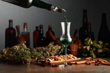 Yeşil bitkisel likör eski bir şişeden bardağa dökülür. Kurutulmuş otlar, çiçekler, baharatlar ve eski mutfak aletleri. Bitkisel ilaç kavramı.