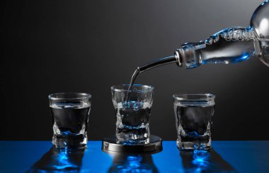 Votka şişeden koyu bir arka planda bir bardağa dökülüyor. Mavi arka ışık. Boşluğu kopyala.