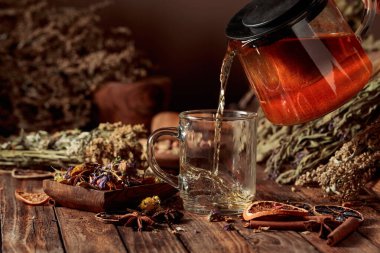 Sıcak bitki çayı cam bardağa dökülür. Çeşitli kurutulmuş otlar ve baharatlar eski bir ahşap masaya serilmiştir. Aromaterapi ya da bitkisel tıp konsepti.