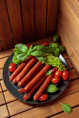 Siyah tabakta ince tütsülenmiş Polonya sosisi. Tahta bir masada fesleğen, salata ve domatesli sosisler..