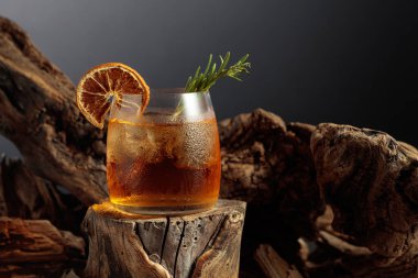 Eski moda bir kokteyl. Eski bir kütüğün üzerinde buz var. Kurutulmuş portakal dilimi ve biberiye dalıyla süslenmiş soğuk, ferahlatıcı bir içecek..