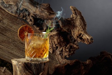 Eski usul bir kokteyl, buzlu, tahta bir kütükte. Kurutulmuş portakal dilimi ve yanan biberiye filizi ile süslenmiş soğuk, ferahlatıcı bir içecek..