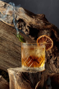 Eski usul bir kokteyl, buzlu, tahta bir kütükte. Kurutulmuş portakal dilimi ve yanan biberiye filizi ile süslenmiş soğuk, ferahlatıcı bir içecek..