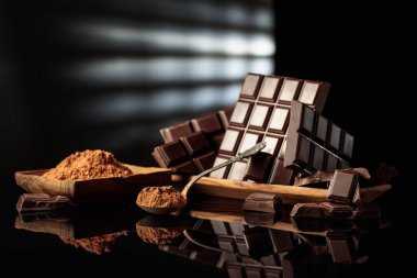 Kırık çikolatalar ve kakao tozlu ahşap tabaklar. Siyah parlak arka plan derinlik katıyor. Bir kaşığa odaklan. Boşluğu kopyala.