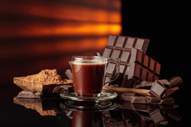 Bir kupa sıcak çikolataya karşı kırık çikolata ve kakao tozlu ahşap tabak. Siyah parlak arka plan derinlik katıyor. Boşluğu kopyala.