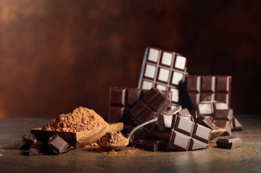 Tahta bir kase kakao tozunun yanında koyu çikolata kırıntıları. Sıcak kahverengi arka plan çikolata renginin zenginliğini vurgular. Boşluğu kopyala.