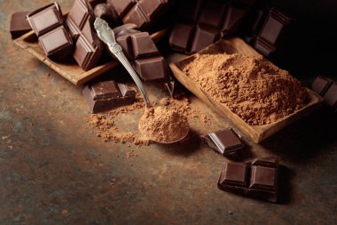 Tahta bir kase kakao tozunun yanında koyu çikolata kırıntıları. Sıcak kahverengi arka plan çikolata renginin zenginliğini vurgular. Bir kaşığa odaklan..