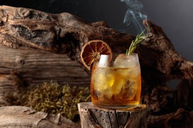 Eski usul bir kokteyl, buzlu, tahta bir kütükte. Kurutulmuş portakal dilimi ve yanan biberiye filizi ile süslenmiş soğuk, ferahlatıcı bir içecek..