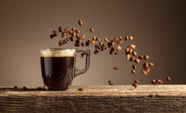 Eski ahşap bir masada cam bardakta espresso. Kavrulmuş kahve çekirdekleri hareket halinde. Boşluğu kopyala.