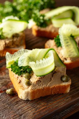 Eski bir kesme tahtasında ezmeli tost. Pate, taze salatalık, kapari ve maydanozlu sandviç..