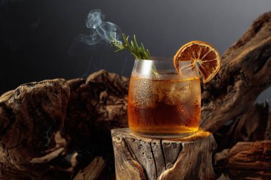 Eski usul bir kokteyl, buzlu, tahta bir kütükte. Kurutulmuş portakal dilimi ve yanan biberiye filizi ile süslenmiş soğuk, ferahlatıcı bir içecek..