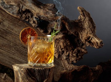 Eski usul bir kokteyl, buzlu, tahta bir kütükte. Kurutulmuş portakal dilimi ve yanan biberiye filizi ile süslenmiş soğuk, ferahlatıcı bir içecek..