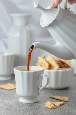 Beyaz porselen fincanda sade kahve ve gevrek kare kraker. Kahve demlikten fincana doldurulur. Gri taş masa ve beyaz çatal bıçaklar.