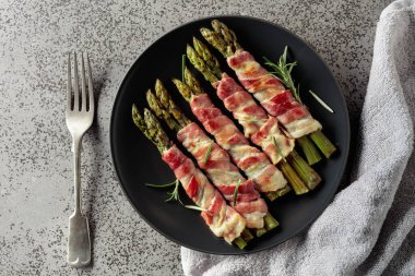 Domuz pastırmasına sarılı ızgara yeşil kuşkonmaz. Fotokopi alanı olan gri taşlı arka planda siyah tabakta ev yapımı aperatif. Üst görünüm.