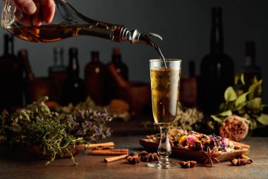 Sarı bitkisel likör ya da karışım eski bir şişeden bardağa dökülür. Kurutulmuş otlar, çiçekler, baharatlar ve eski mutfak aletleri. Bitkisel ilaç kavramı.