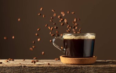 Eski bir tahtanın üzerinde cam bardakta sade kahve. Kavrulmuş kahve çekirdekleri hareket halinde. Boşluğu kopyala.